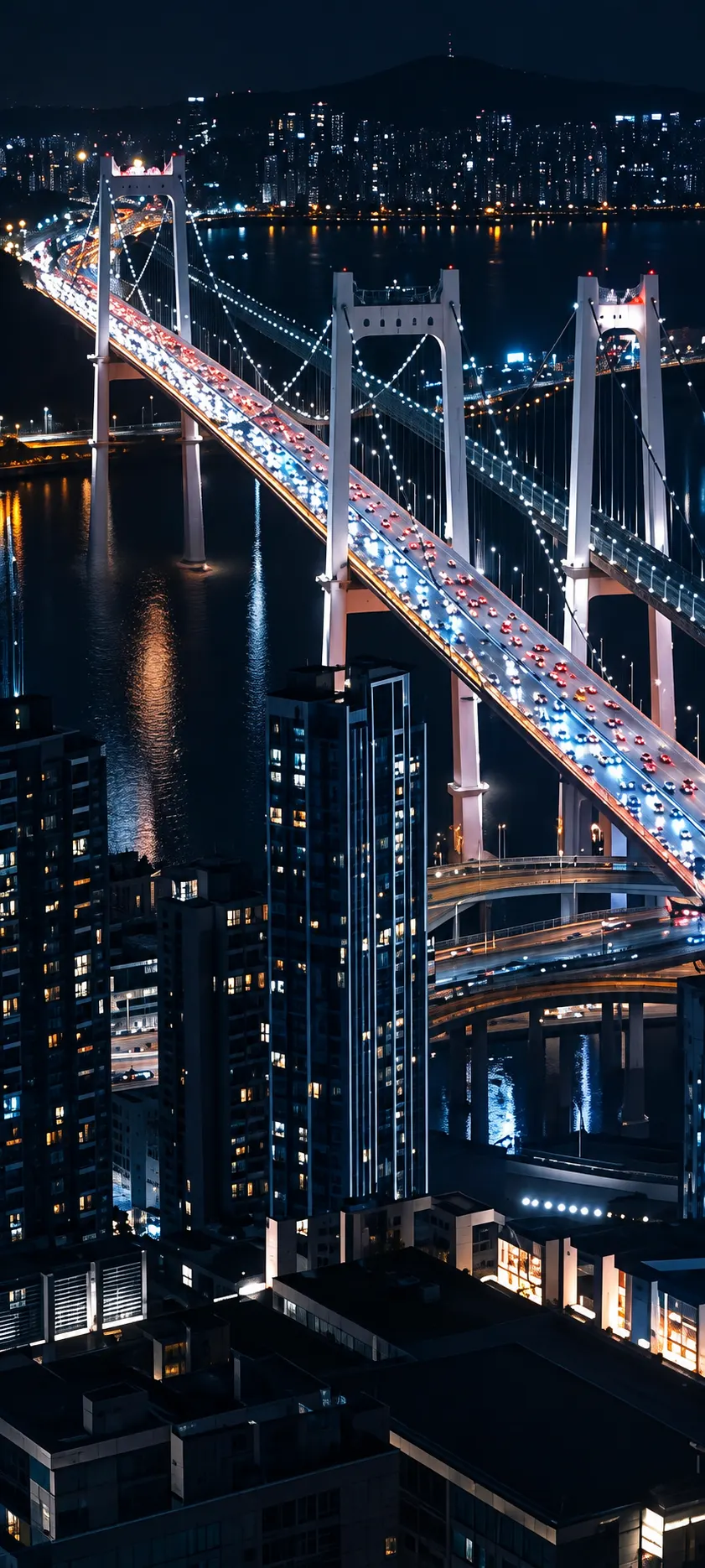 A lit suspension bridge crossing a harbor beside a city skyline at night